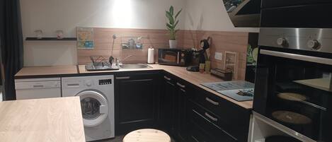 Cocina compacta con lavadora, lavaplatos, microondas y estufa. Hay dos taburetes y una mesita.