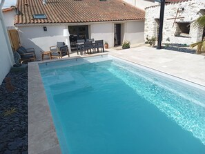 Une piscine rectangulaire, bordée d’une terrasse carrelée, entourée de mobilier d’extérieur et d’un mur en pierre.