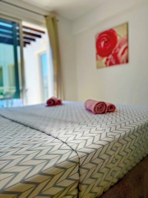Ein Hotelzimmer mit einem Bett, das eine geometrische Bettdecke, ein aufgerolltes Handtuch auf dem Bett und ein Gemälde einer roten Rose an der Wand zeigt.
