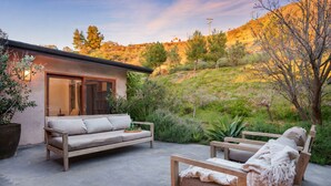 Une terrasse avec un canapé et un fauteuil, entourée de verdure et adossée à une colline.