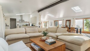 Un salon spacieux avec un grand canapé d’angle, une table basse en bois sur laquelle sont posés des livres, et une vue sur la cuisine et la salle à manger grâce à de grandes fenêtres.