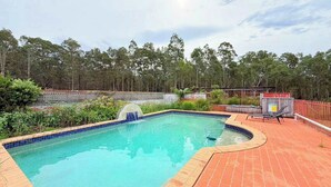 A rectangular swimming pool with a waterfall feature, surrounded by a brick deck and a wooden fence. - Affordable Queen Room in Large Farmhouse (Worrigee)