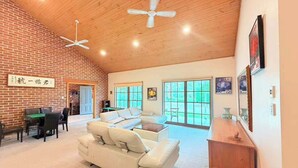 A spacious living room with a brick wall, a white sofa, a wooden coffee table, and a ceiling fan. - 1BR Near Bus Stop with Pool & Farm Views (Worrigee)