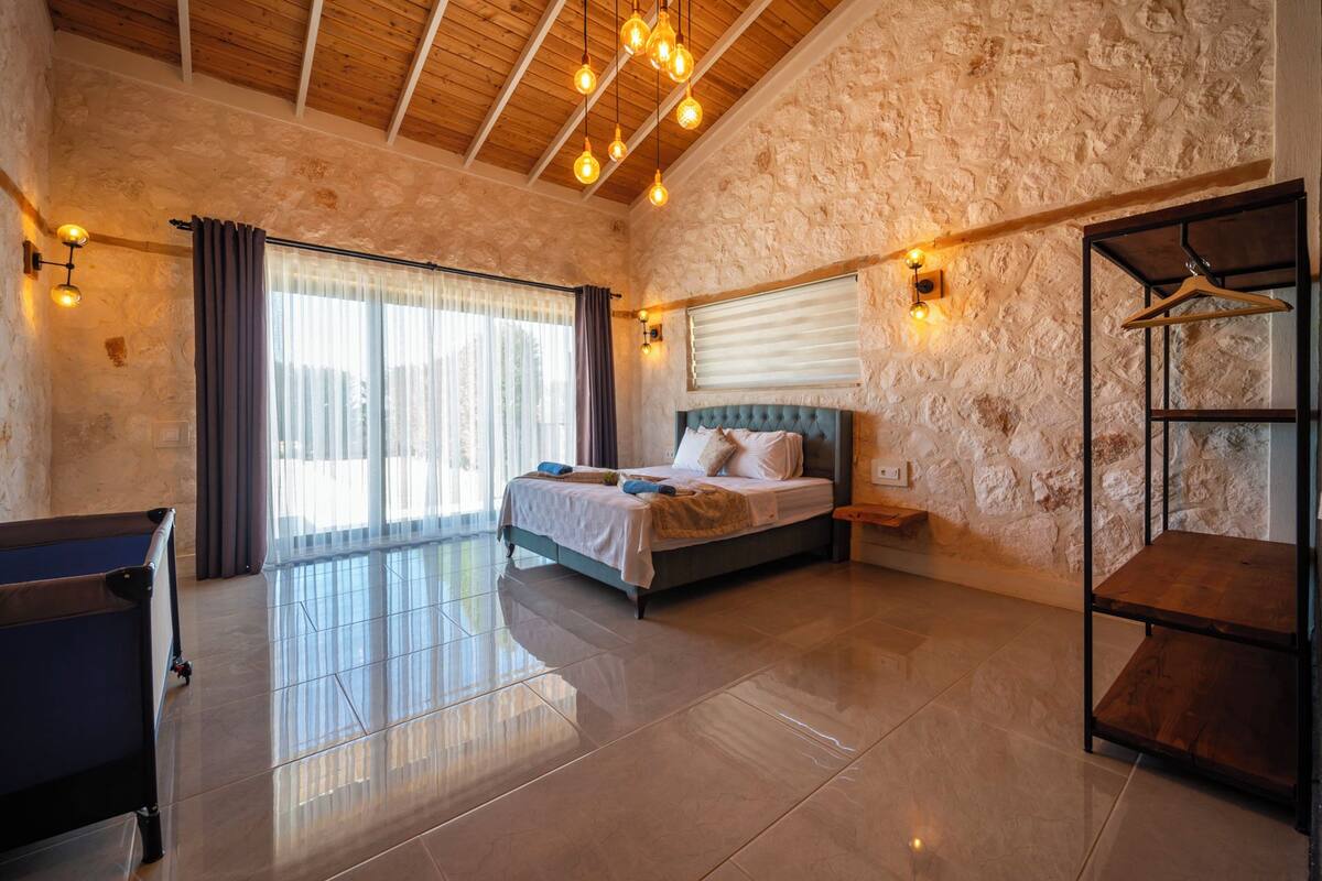A bedroom with a bed, a wooden ceiling, stone walls, a window with curtains, and a wooden shelf.