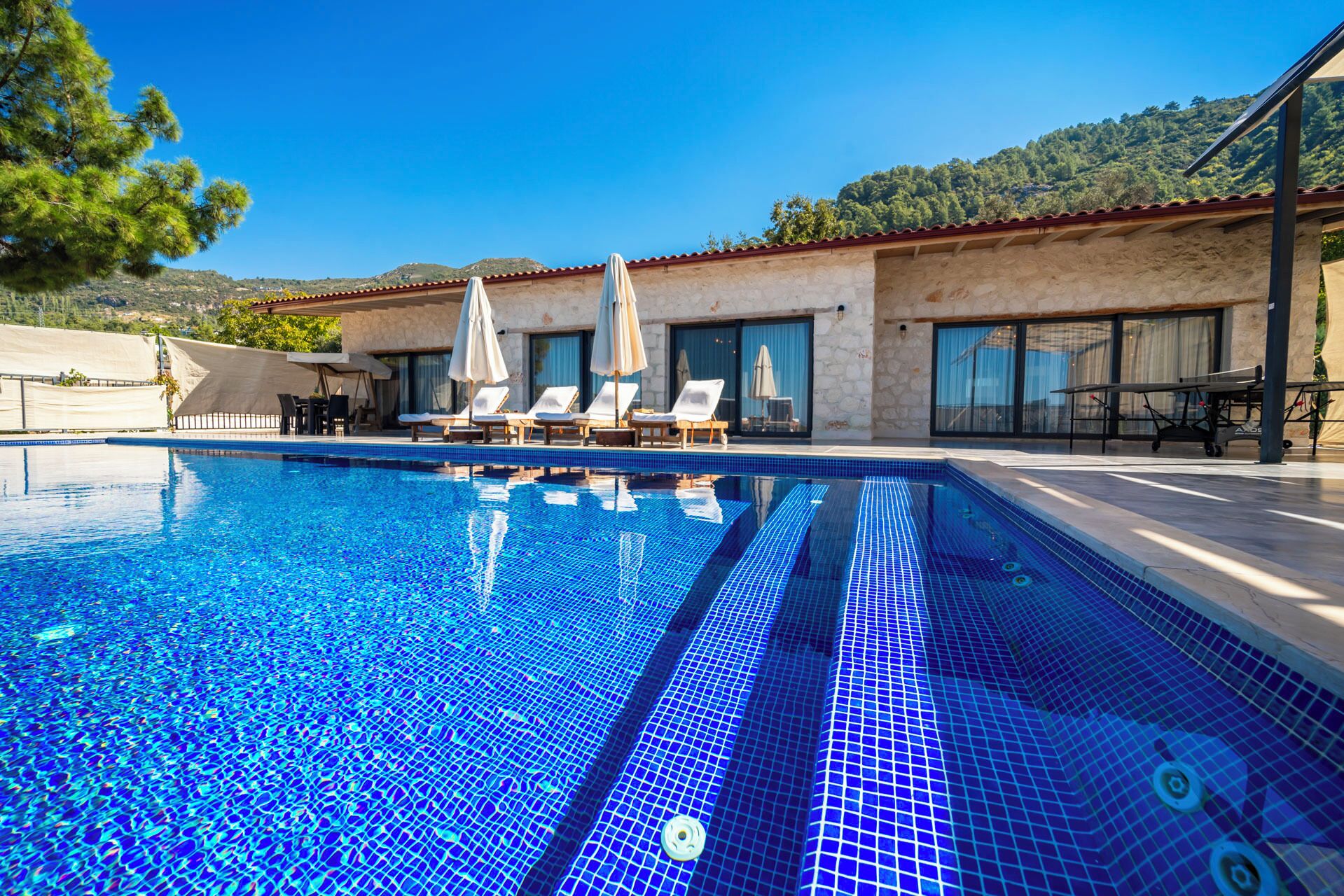 A swimming pool with clear blue water, surrounded by lounge chairs and umbrellas, with a mountain backdrop.