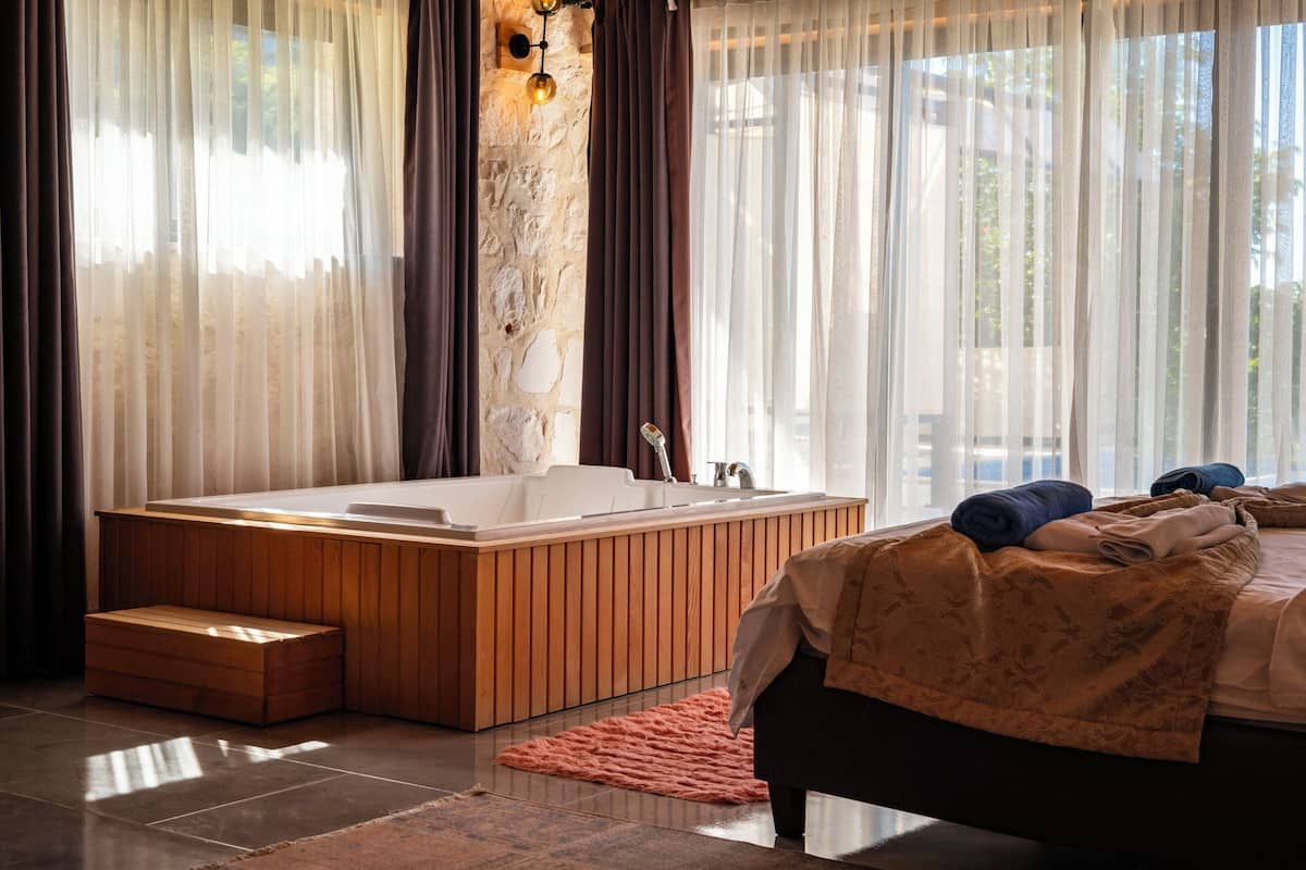 A modern bathroom with a wooden bathtub, a large window with sheer curtains, and a stone wall.