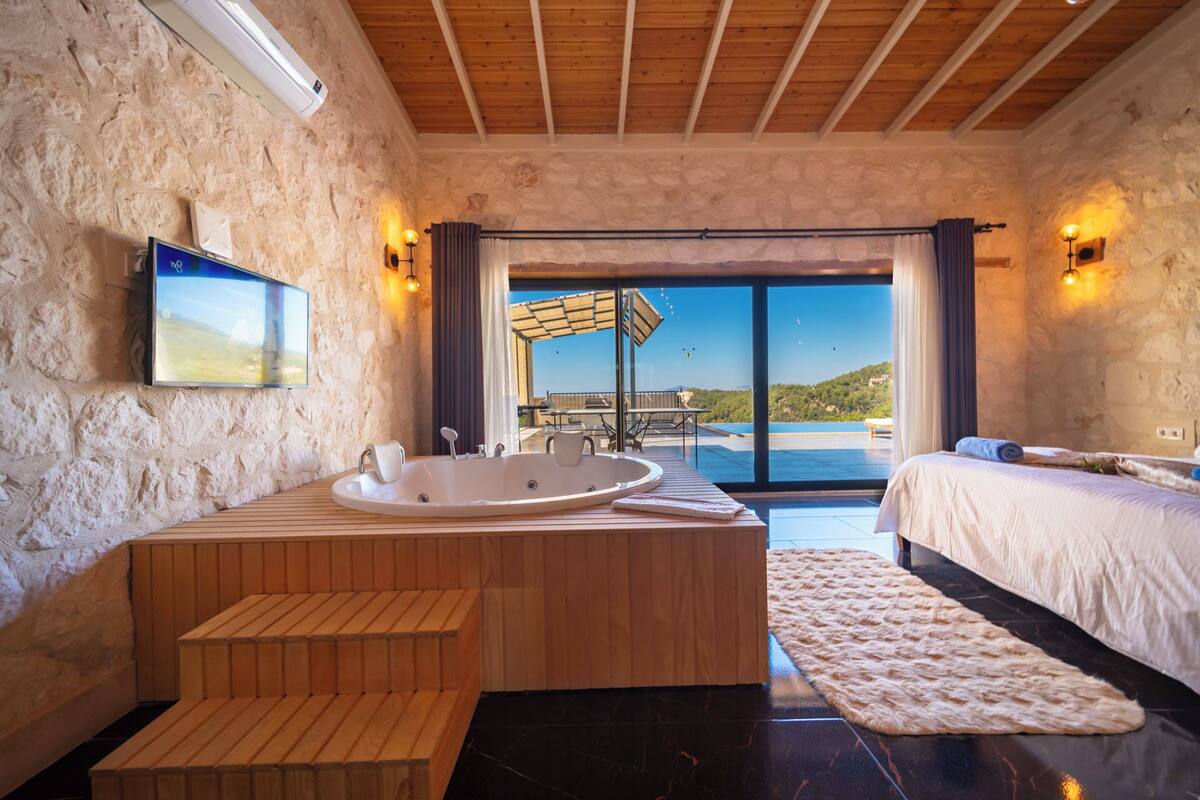 A modern bathroom with a large bathtub, wooden bench, and a view of a pool and hills.