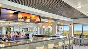 A bar area with a long counter, multiple hanging light fixtures, and a large TV screen displaying a sunset scene. - CT 7616 Golf Course View - Welcome! (Fort Pierce)