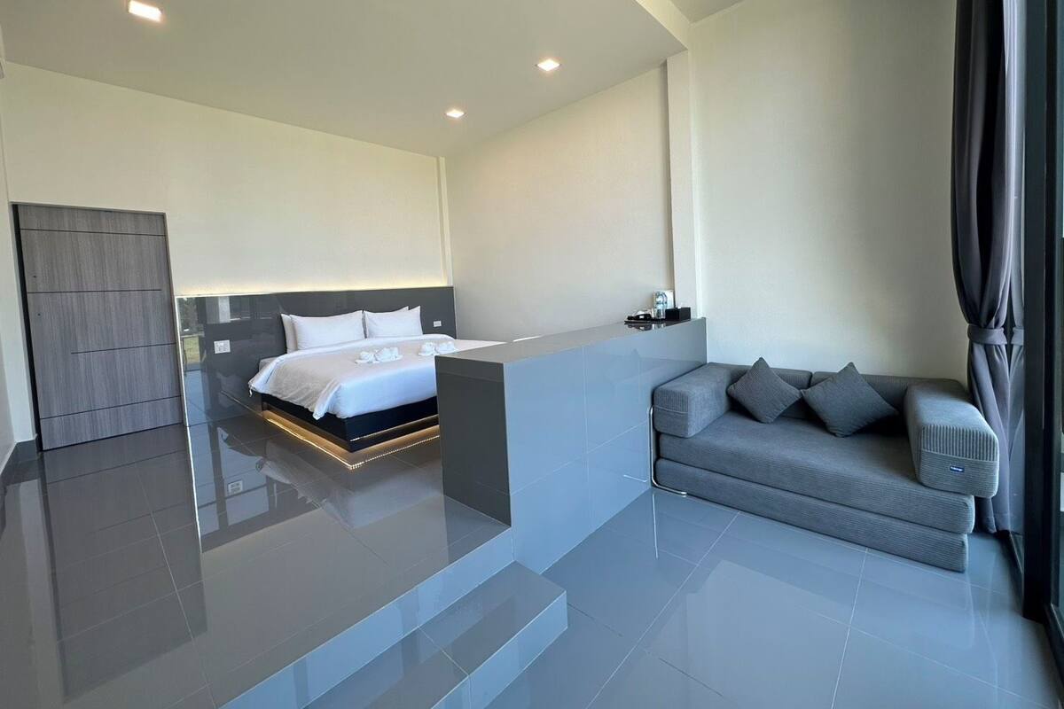 A modern hotel room with a large bed, a grey sofa, and a small table with a lamp.