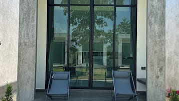 Un edificio moderno con un amplio ventanal, dos sillones de descanso y una zona de patio con baldosas.