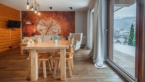 Ein moderner Essbereich mit einem Holztisch und Stühlen, einem großen Fenster mit Blick auf verschneite Berge und einer Wand mit einer großformatigen Holzmaserung-Kunst.