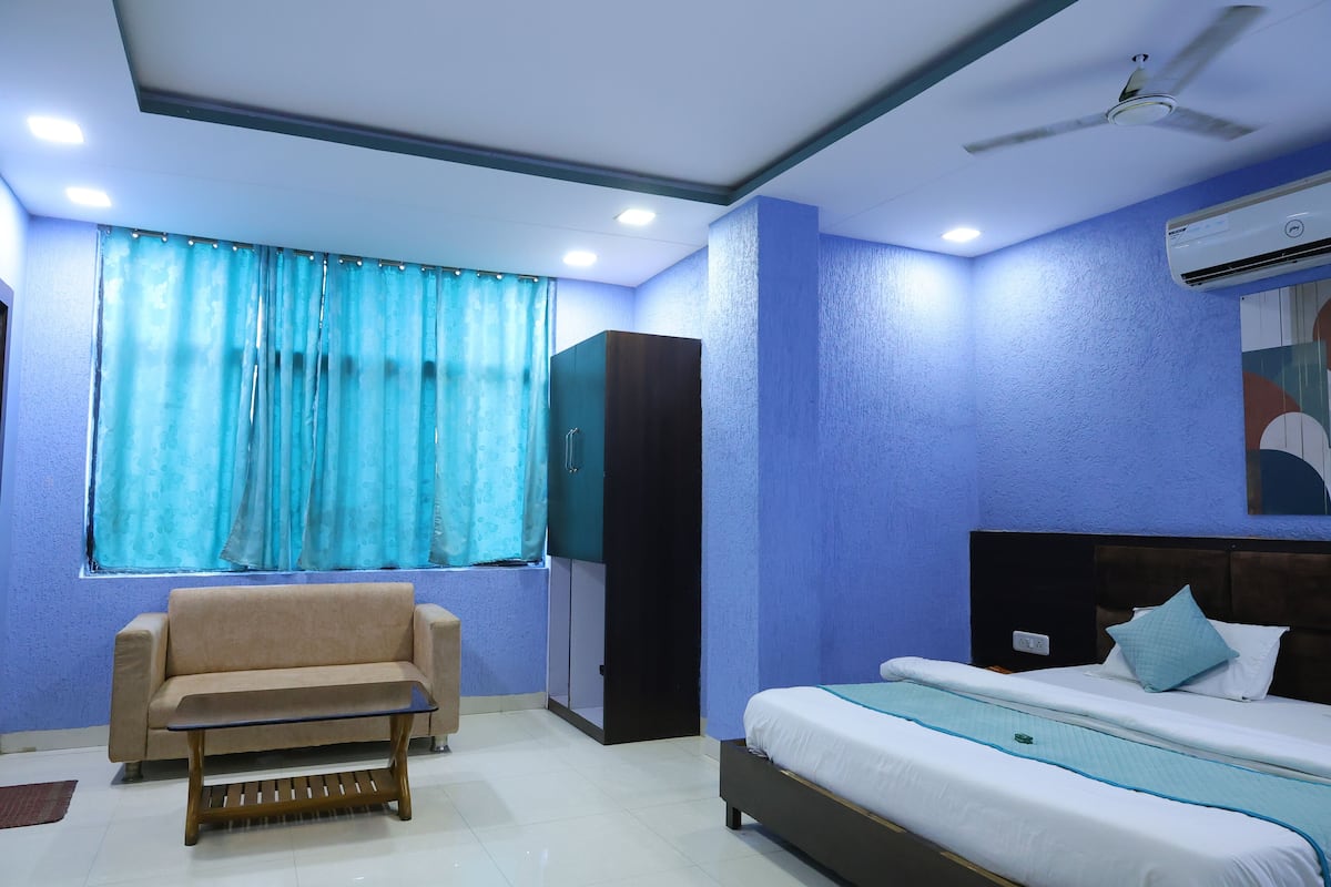 A room with a bed, a sofa, a small table, a window with curtains, a ceiling fan, and an air conditioning unit.