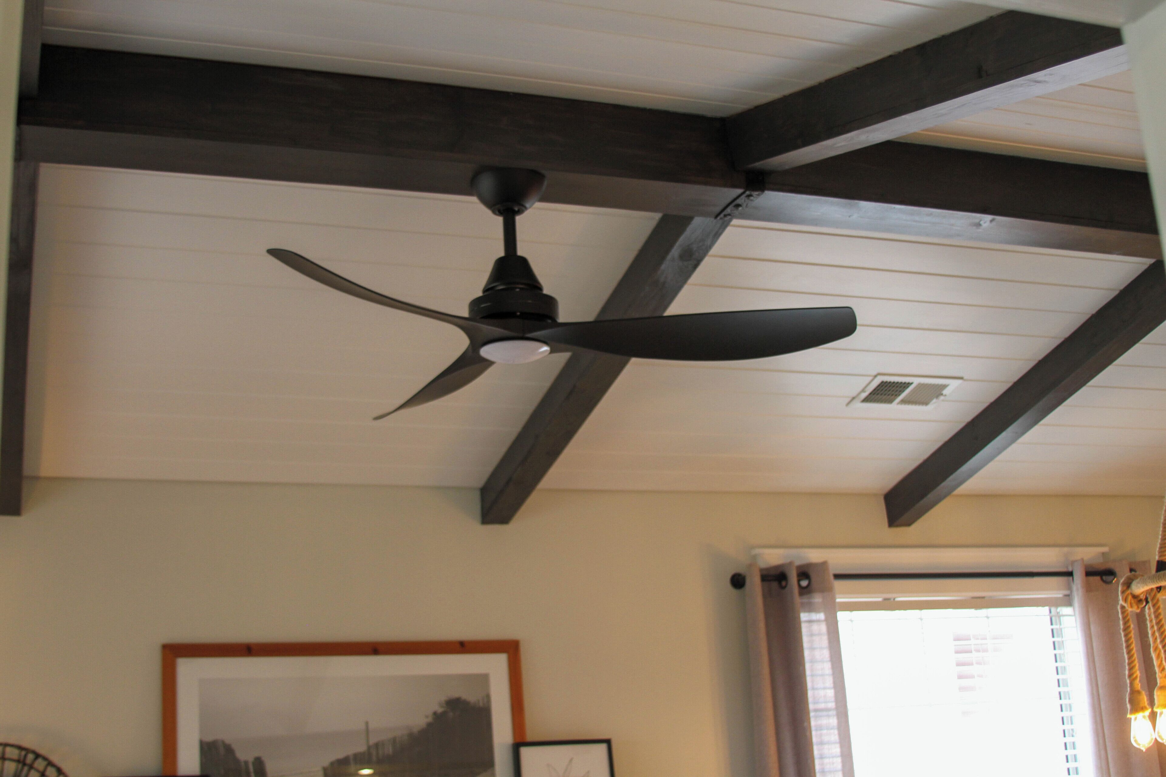 A ceiling fan in a room with wooden beams, a framed picture on the wall, and a window with curtains.