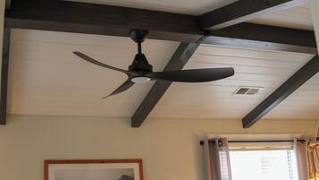 A ceiling fan in a room with wooden beams, a framed picture on the wall, and a window with curtains.