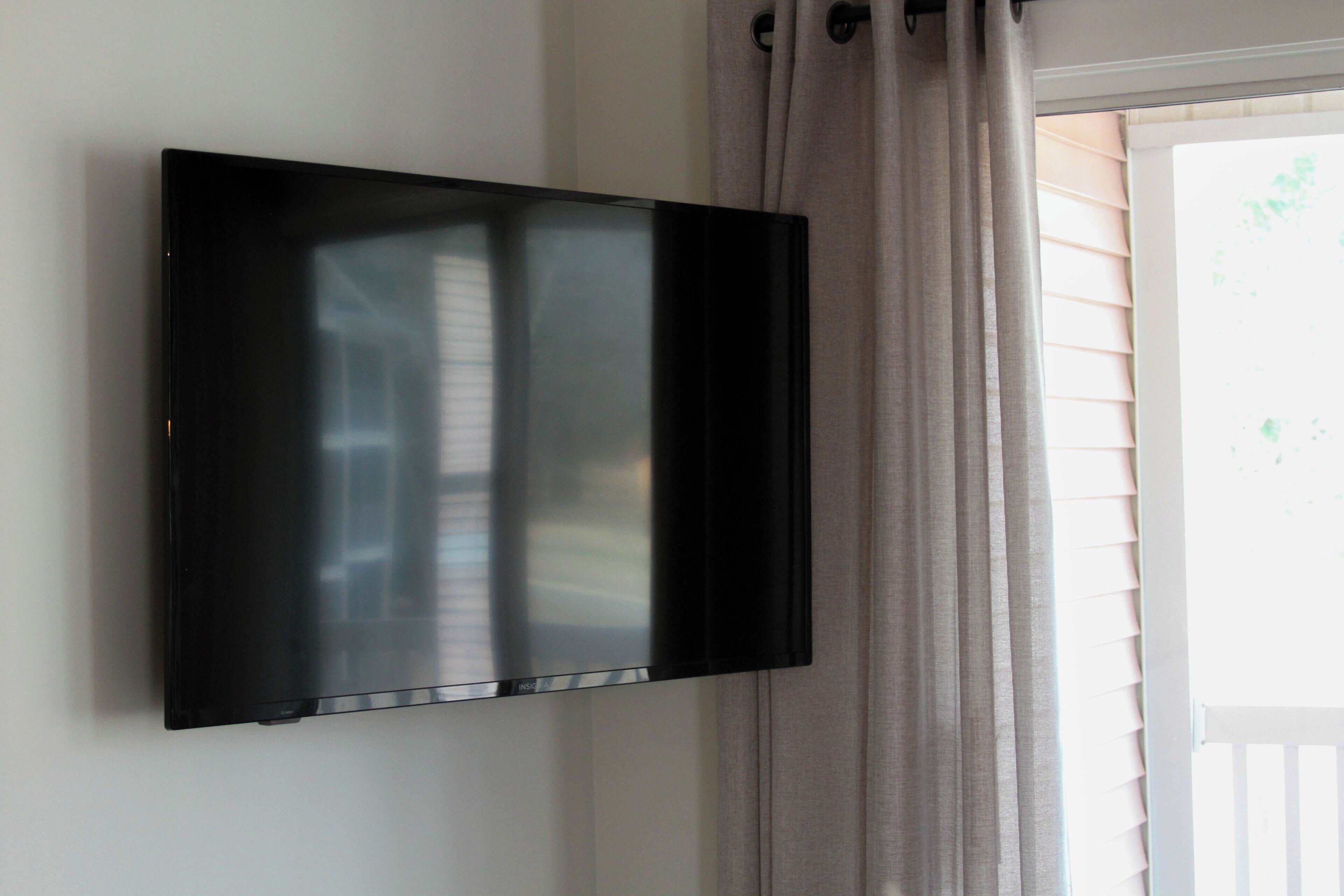 A flat-screen TV mounted on a wall in a room with a window and blinds.