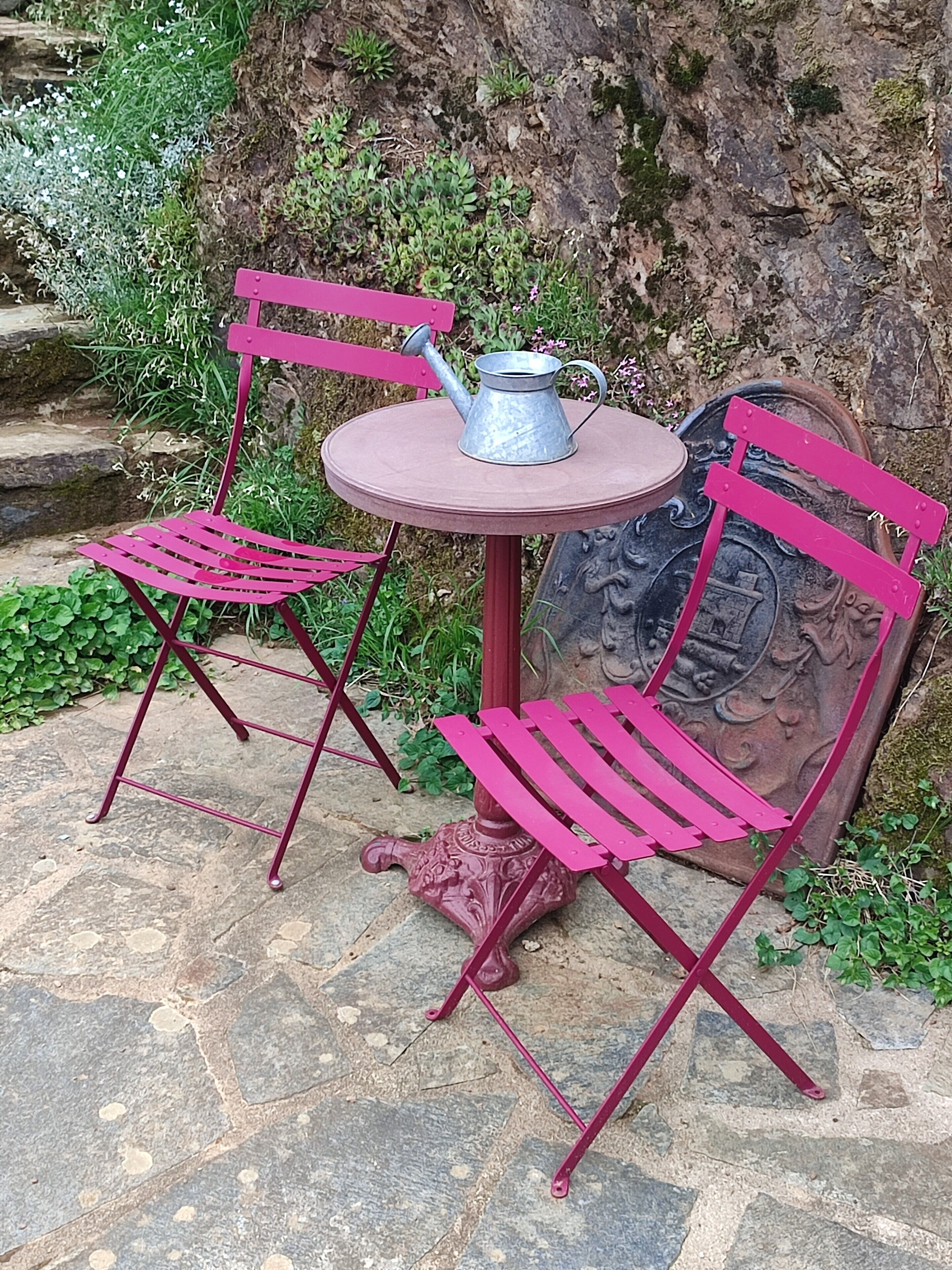 Une table avec un arrosoir en métal et deux chaises roses.