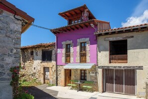 Une rue étroite bordée de bâtiments traditionnels en pierre, dont l’un présente une façade violette et des volets en bois, et l’autre une façade en pierre et une porte en bois.