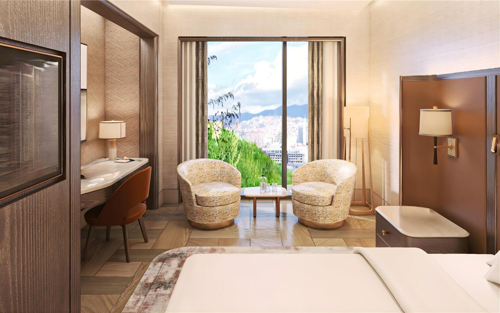 A hotel room with a bed, two armchairs, a desk, and a view of a cityscape through large windows.