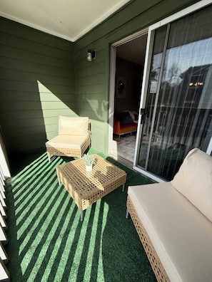 Ein Balkon mit einem grünen Kunstrasenbelag, zwei Korbstühlen, einem kleinen Tisch mit einer Pflanze und einer Couch.