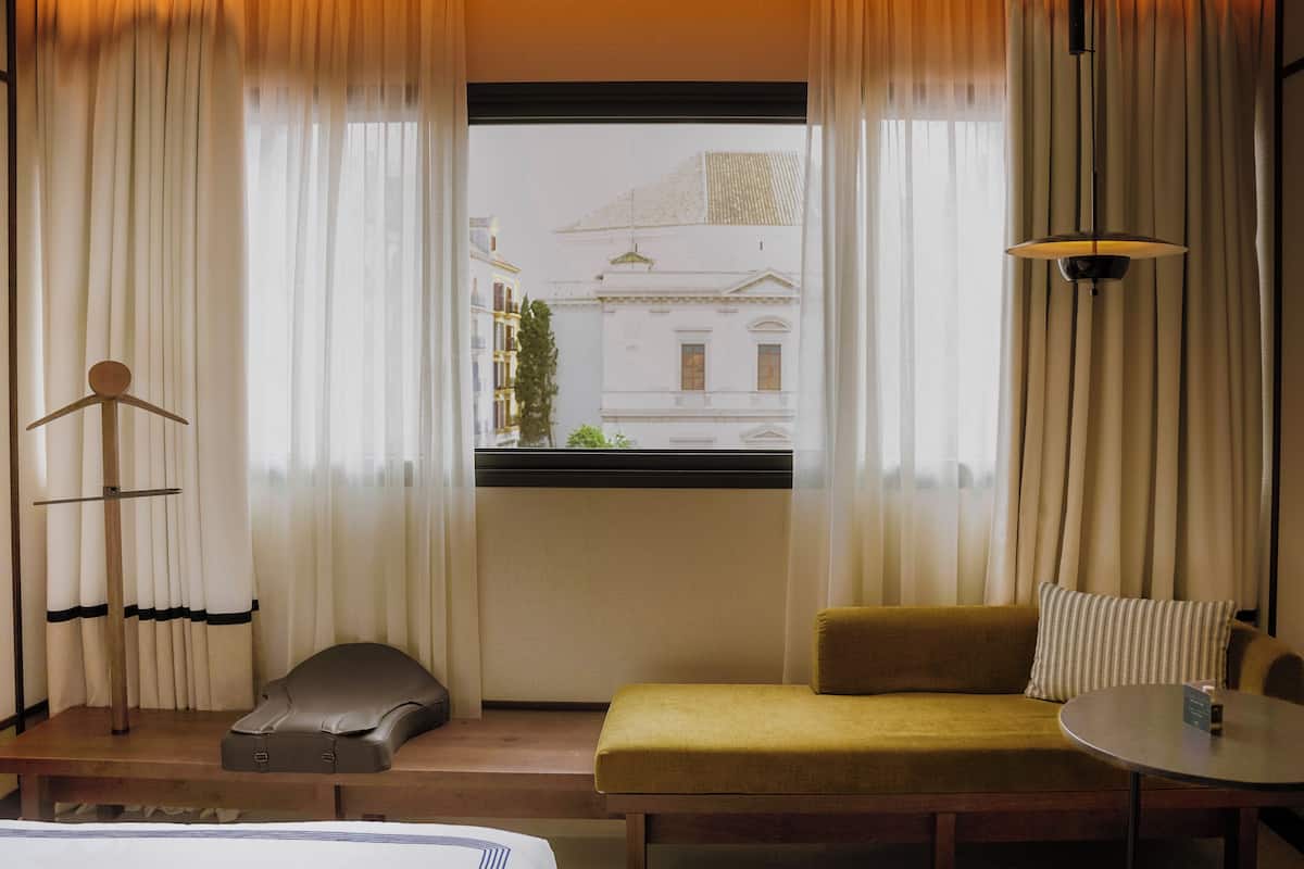 A hotel room with a bed, a sofa, a table, a hanging lamp, and a window with curtains.