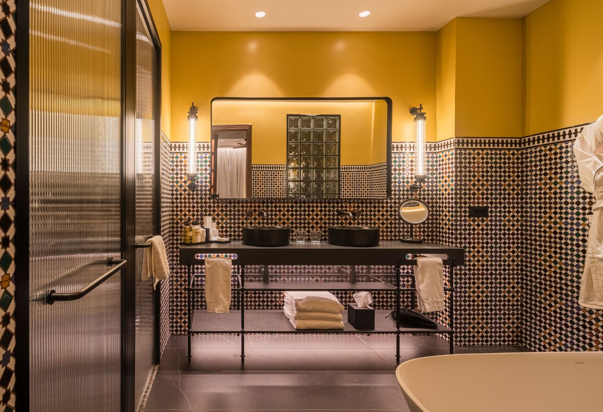 Un baño con una pared amarilla, un salpicadero de azulejos blanco y negro, un espejo, un lavabo con dos cubetas, un perchero para toallas y una bañera.