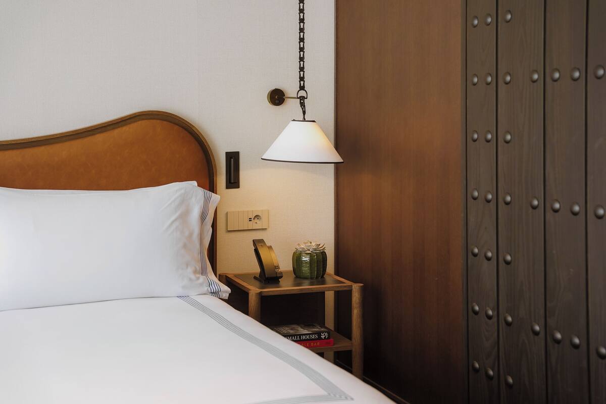 A neatly made bed with white linens, a bedside lamp, a small wooden nightstand with a book and a decorative item, and a wooden wardrobe with a chain light fixture.