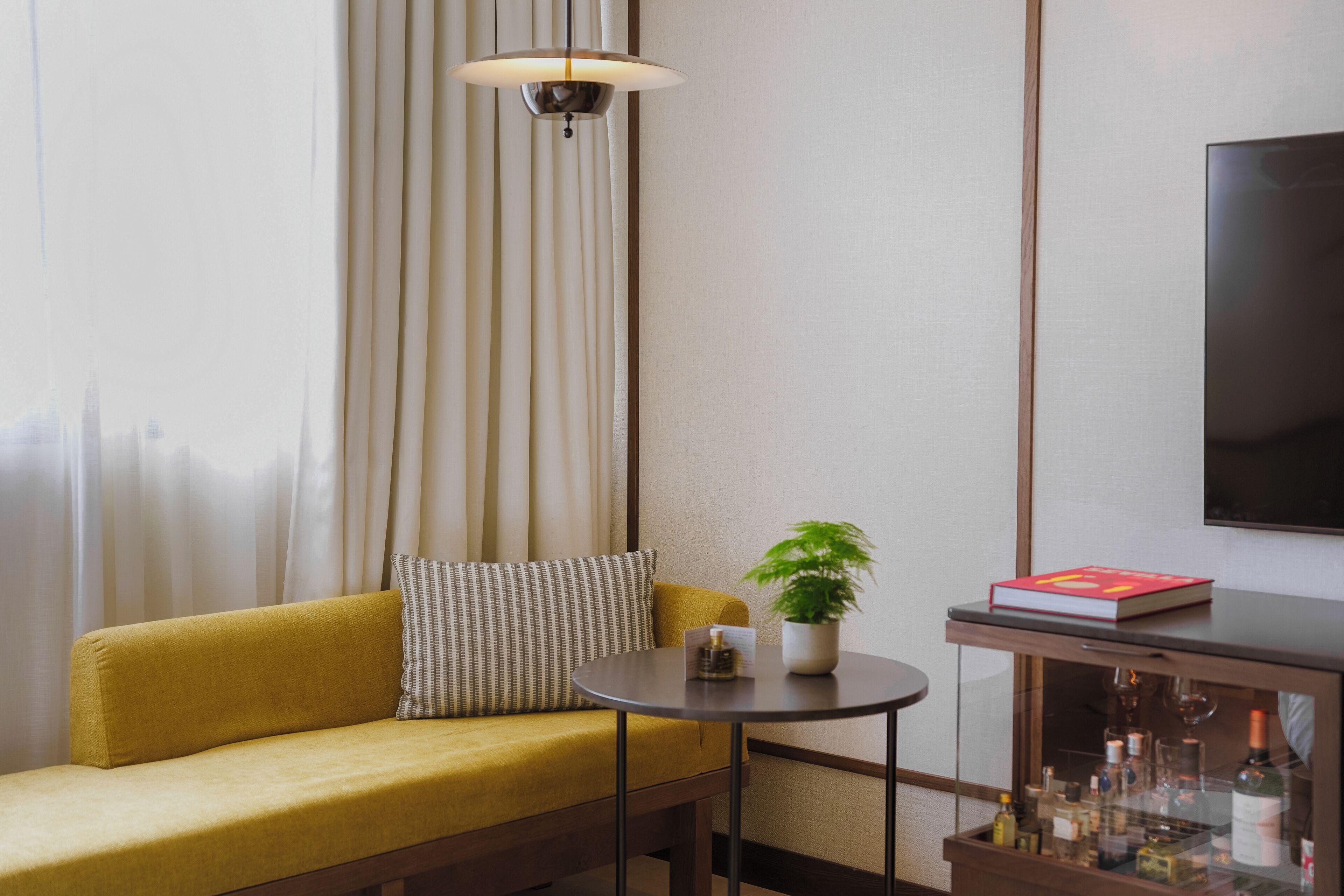A room with a sofa, a small round table with a plant and a book, a cabinet with bottles, and a TV on the wall.