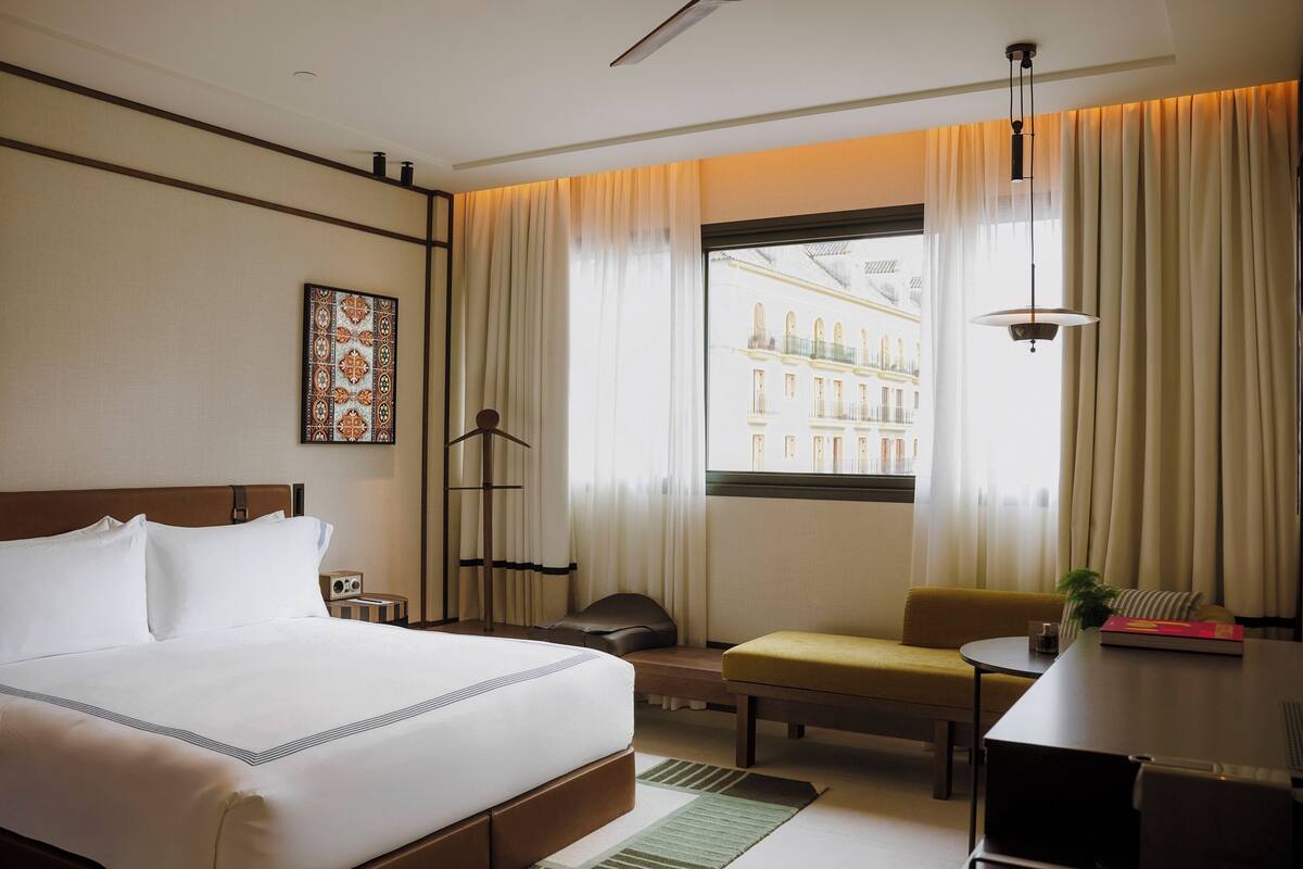 A hotel room with a bed, a desk, a chair, a lamp, and a window with a view of buildings.