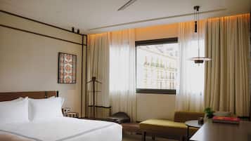 A hotel room with a bed, a desk, a chair, a lamp, and a window with a view of buildings.