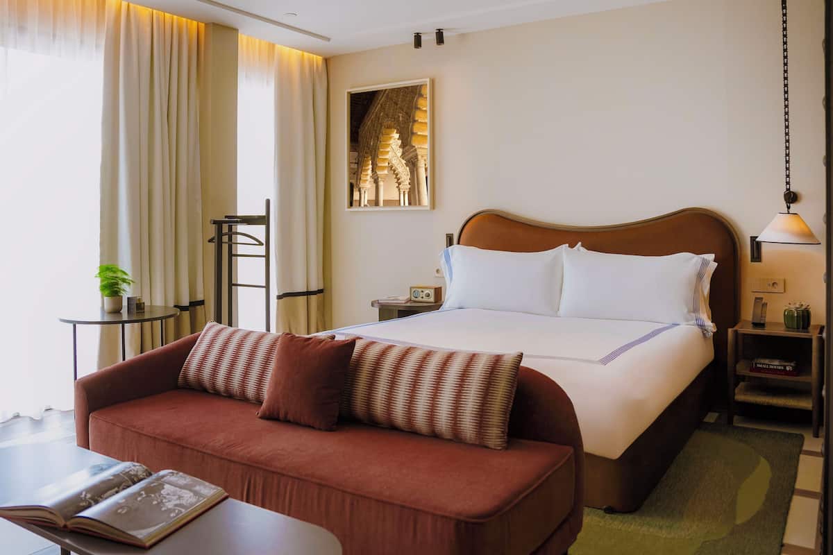 A hotel room with a bed, a sofa, a small table, a lamp, a picture on the wall, and a plant.