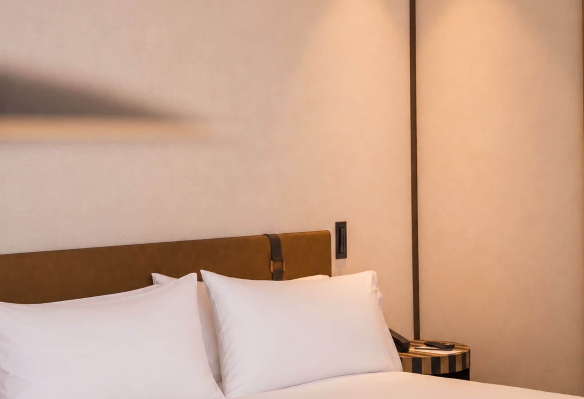 A hotel room with a bed, pillows, a nightstand, and a wall-mounted light fixture.