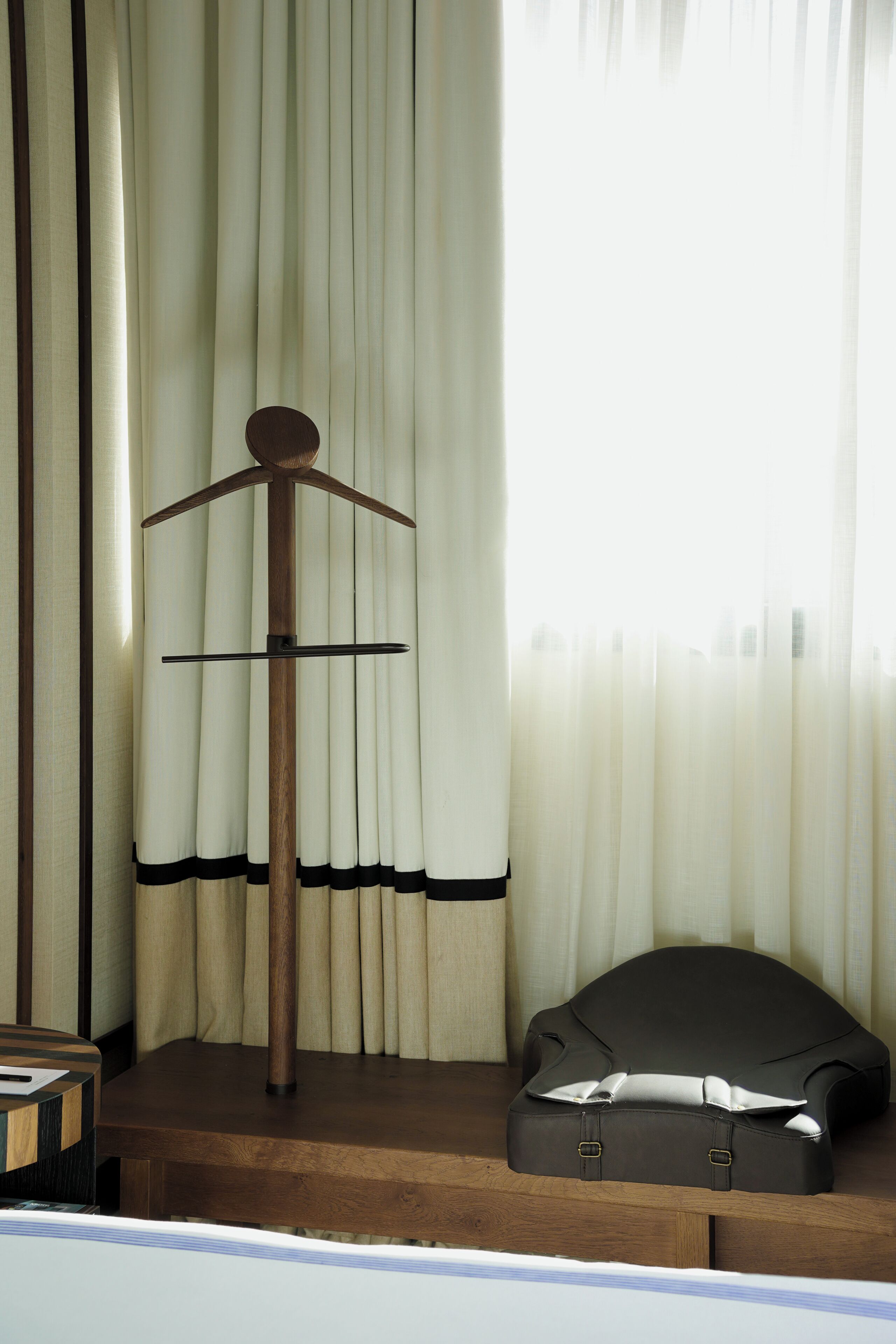 A wooden hanger stand with a coat on it, a suitcase, and a window with curtains.