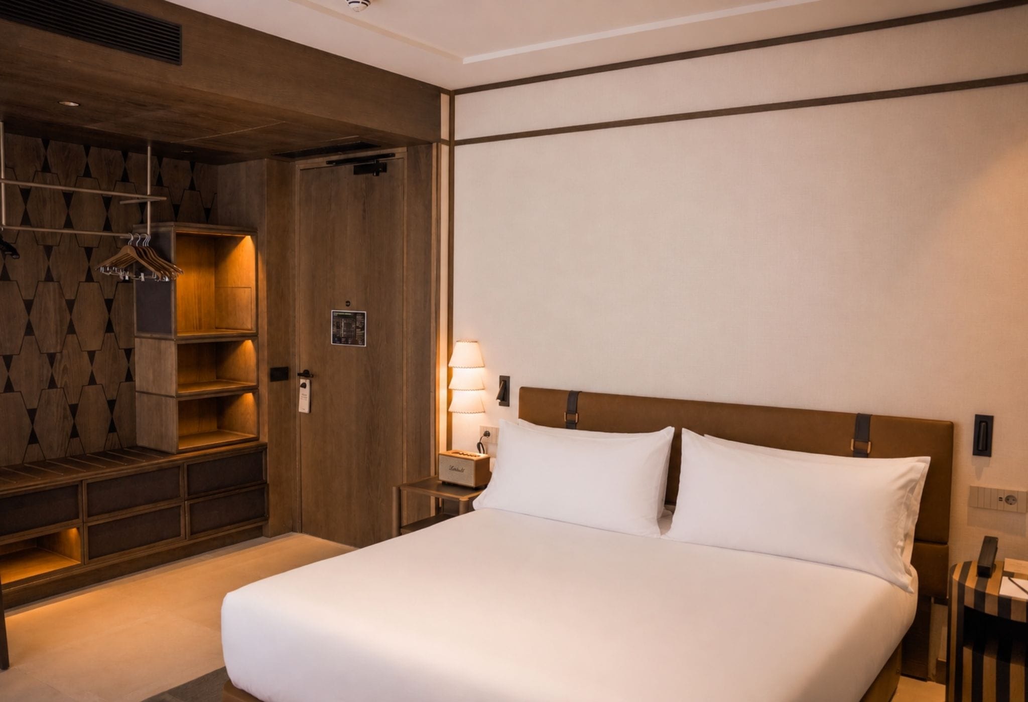 A hotel room with a large bed, wooden shelving, a nightstand, and a wall-mounted lamp.