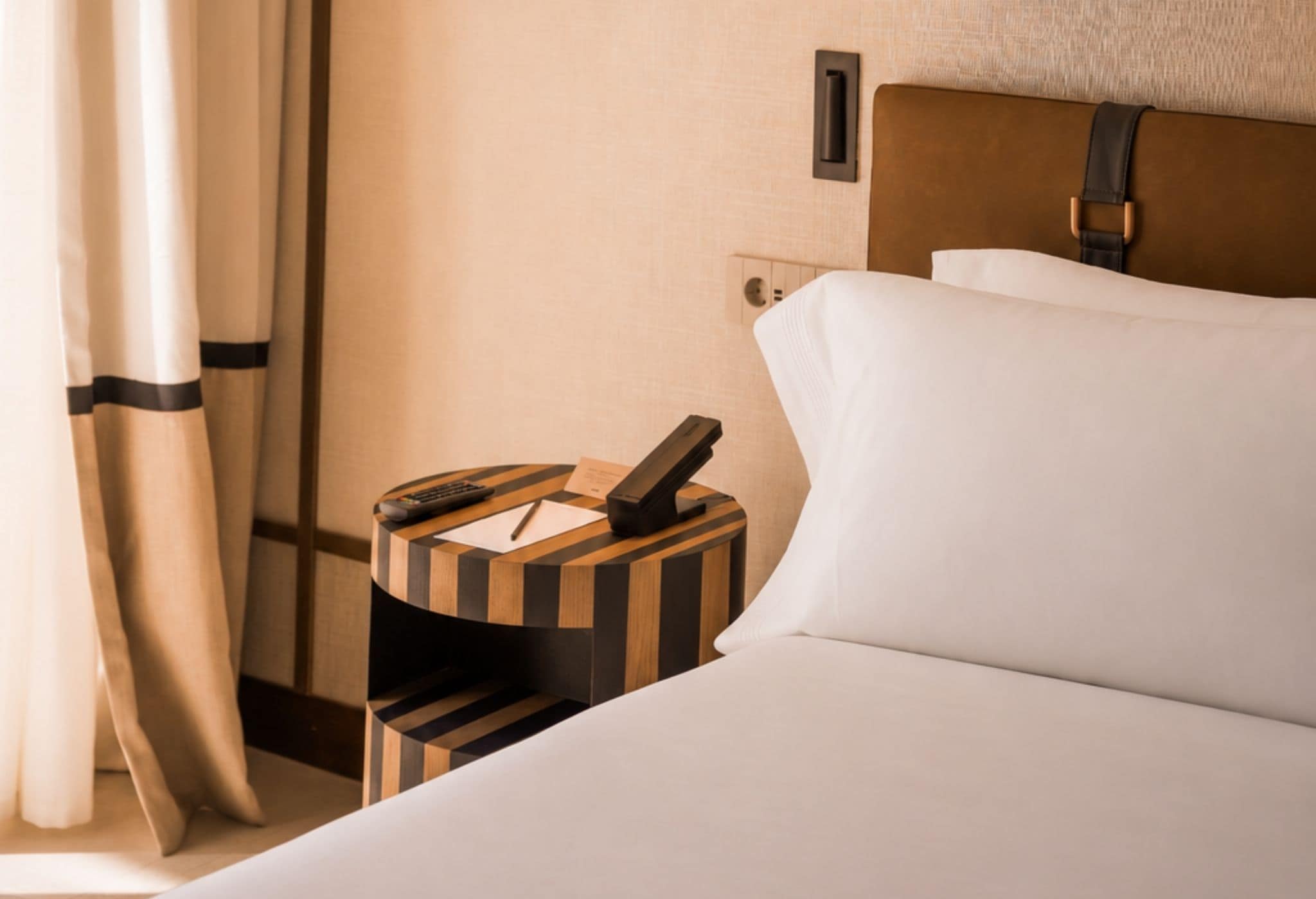 A hotel room with a neatly made bed, a bedside table with a phone and a remote control, and a wall-mounted light switch.