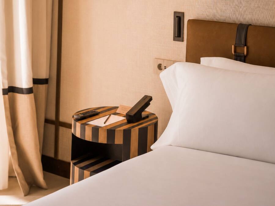 A hotel room with a neatly made bed, a bedside table with a phone and a remote control, and a wall-mounted light switch.