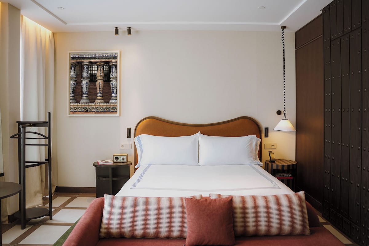 A neatly arranged hotel room with a large bed, a bedside table with a lamp, a framed artwork on the wall, and a wardrobe.