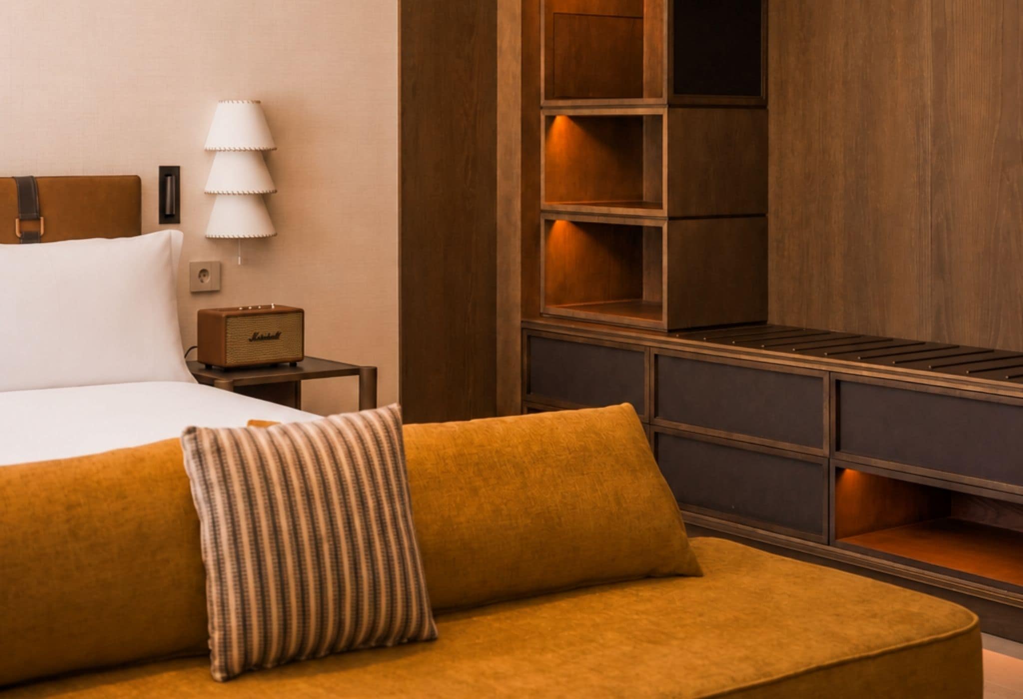 A hotel room with a bed, a bedside lamp, a small table with a radio, a sofa with a striped pillow, and built-in shelving.