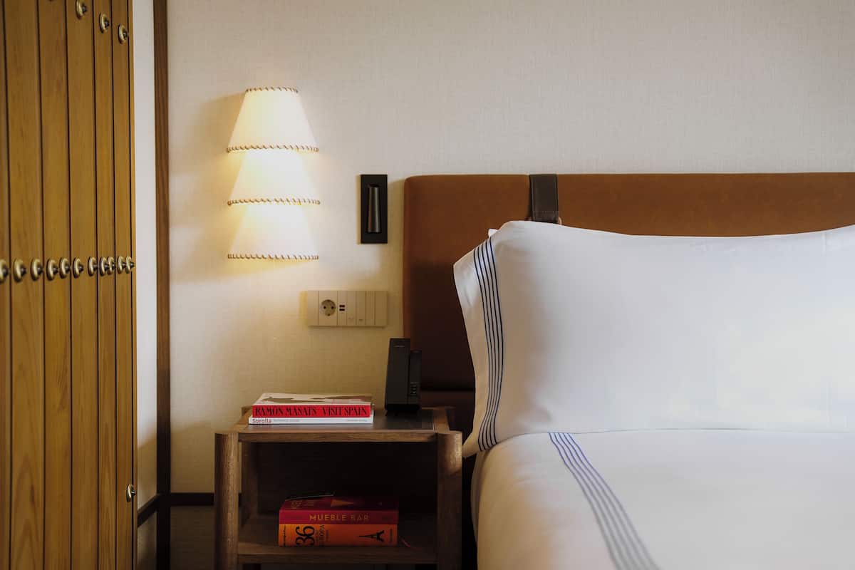 A hotel room with a bed, a nightstand with books, a lamp, and a wall socket.