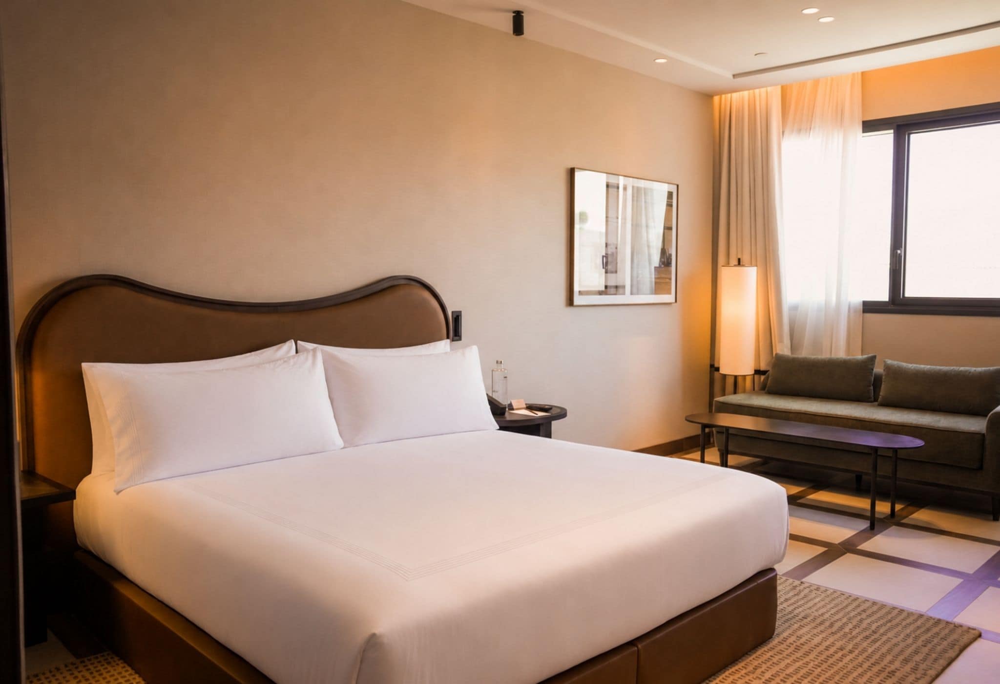 Een hotelkamer met een groot bed, een nachtkastje, een lamp, een bank en een raam met gordijnen.