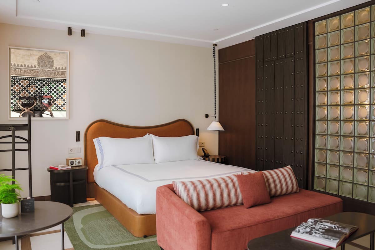 A hotel room with a bed, a sofa, a small table, a lamp, a decorative wall, and a window with a view.