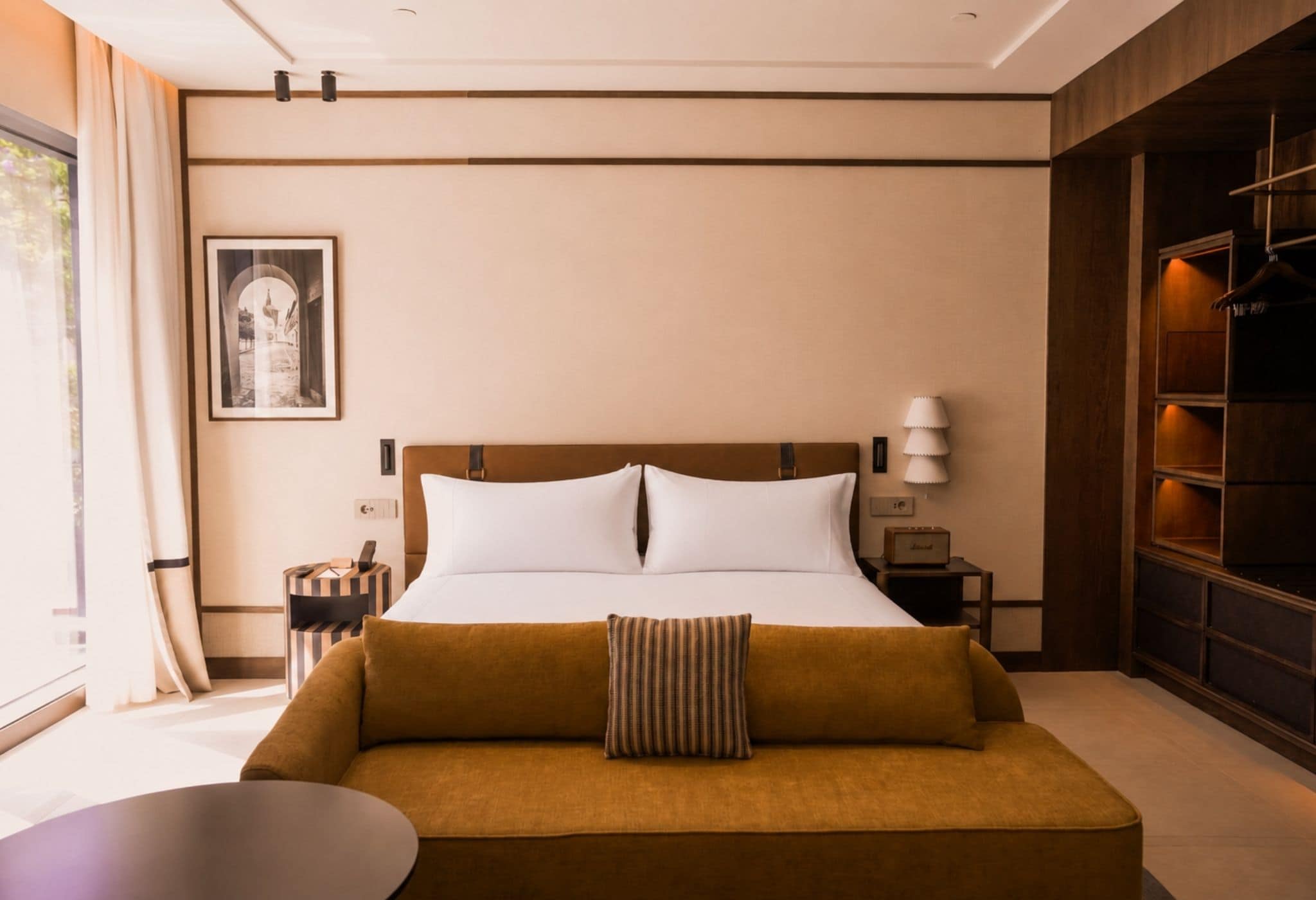 Habitación de hotel con cama, sofá, mesita de noche, lámpara, un cuadro en la pared y una ventana con cortinas.