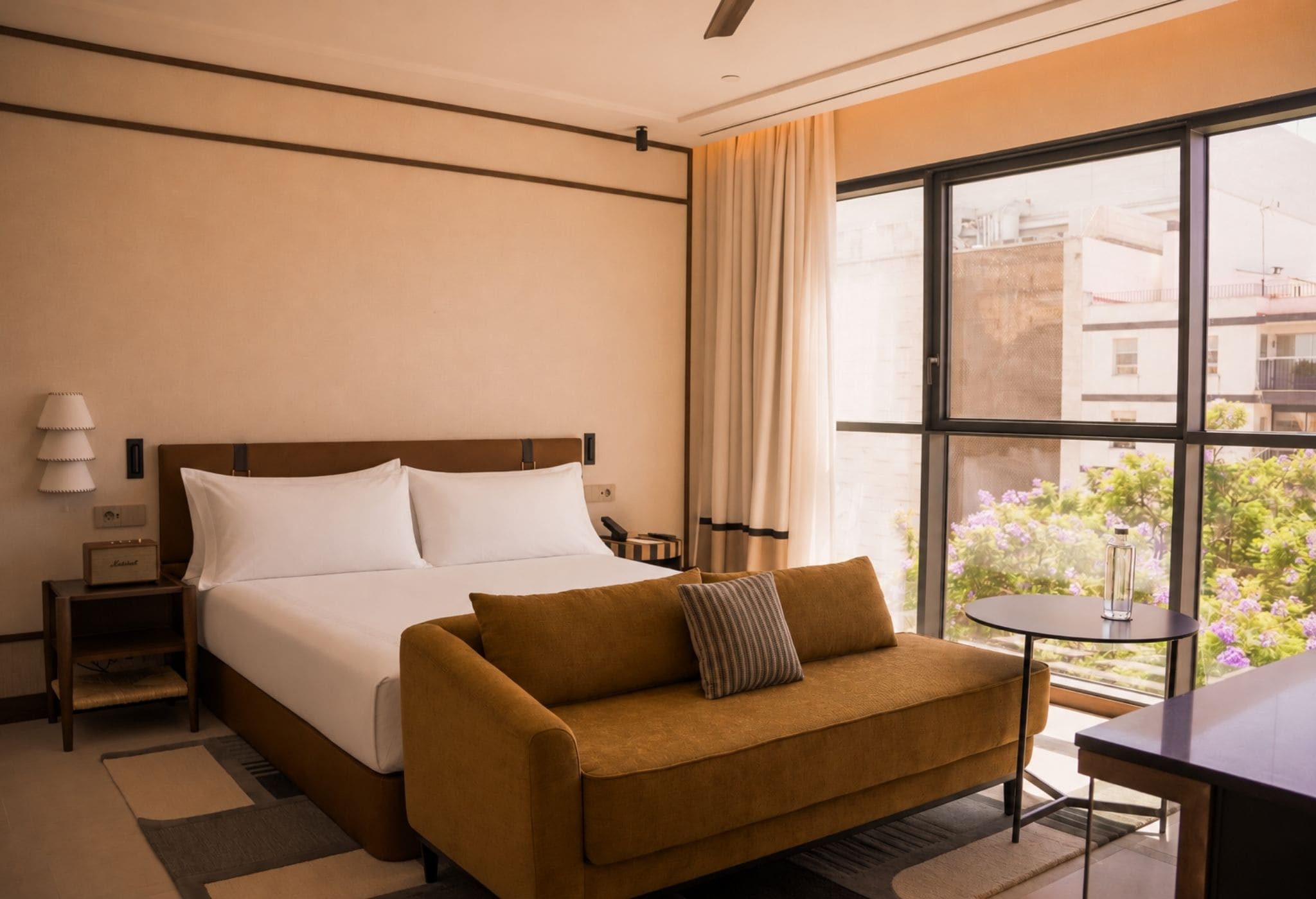Quarto de hotel com cama, sofá, mesa pequena e janela com vista para edifícios e áreas verdes.