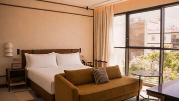 Quarto de hotel com cama, sofá, mesa pequena e janela com vista para edifícios e áreas verdes.