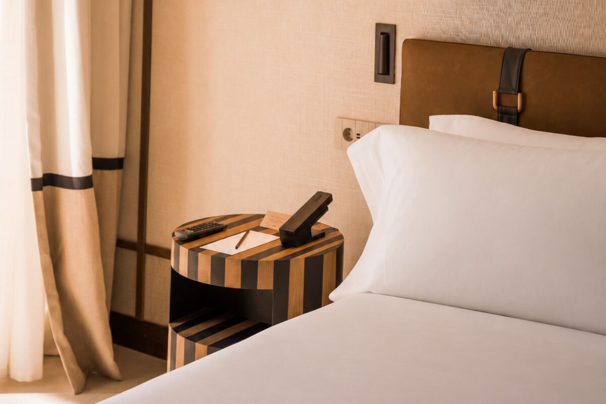 A hotel room with a neatly made bed, a bedside table with a phone and a remote control, and a wall-mounted light switch.