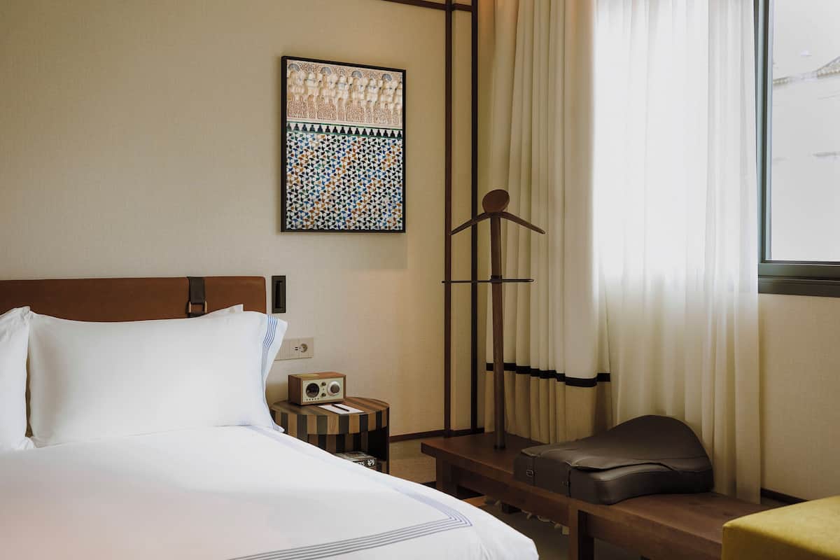 A hotel room with a bed, a bedside table with a radio, a suitcase, and a window with curtains.