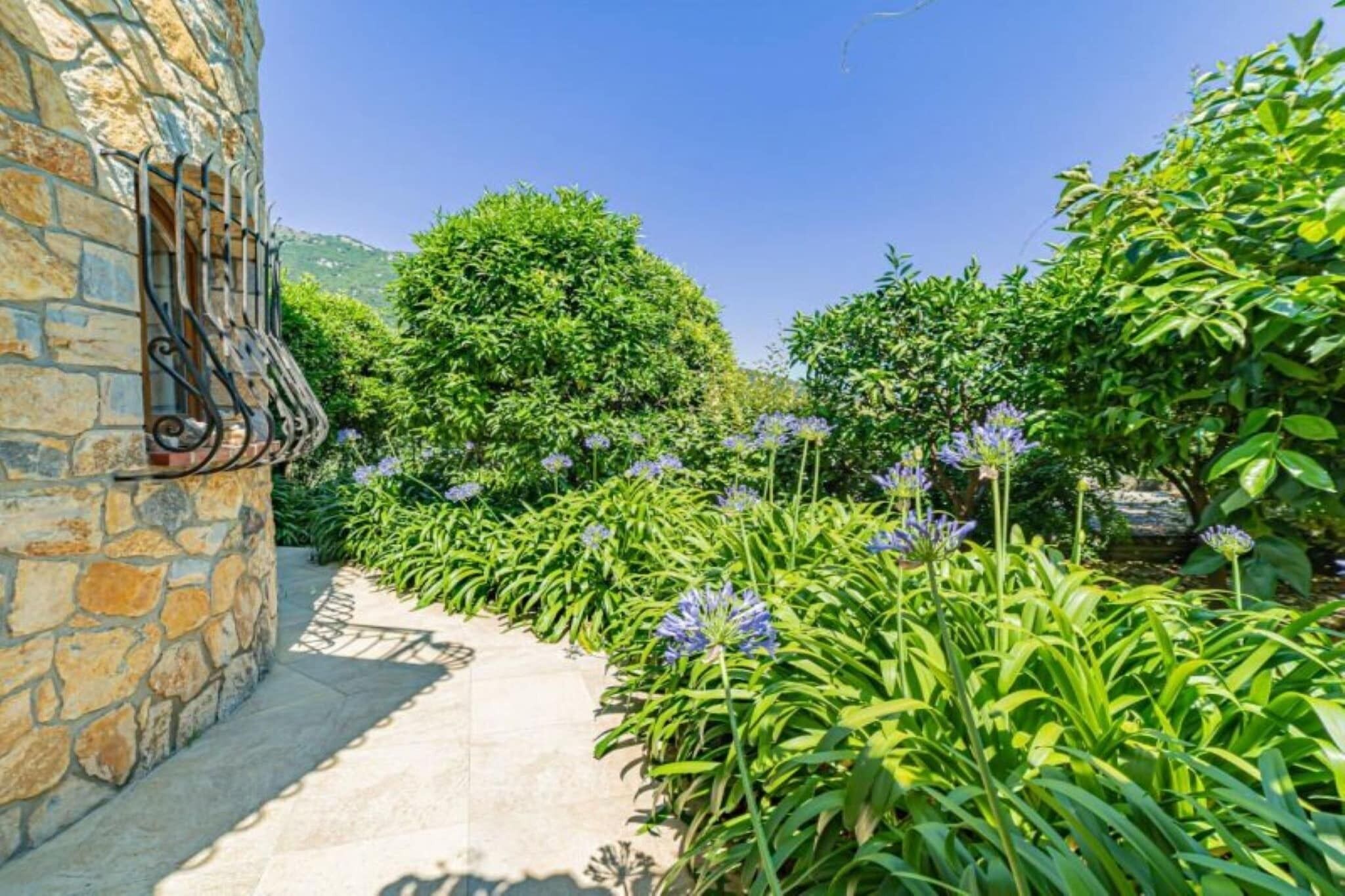 A stone pathway leading to a building with a wrought iron gate, surrounded by lush greenery and flowering plants.