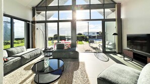 A modern living room with a large glass door leading to a patio, a sofa, a coffee table, and a flat-screen TV. - Architect-designed villa with sea view (Le Conquet)