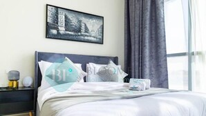 A neatly made bed with white bedding, a framed picture on the wall, and a nightstand with a lamp and a small decorative item. - Bloomfields Magnificent STU in The V (Abu Dhabi)