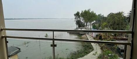 A room with a large window overlooking a body of water, a balcony with a railing, and a view of a distant shoreline with trees and buildings.