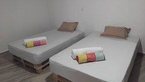 Twee eenpersoonsbedden met geruit kussens en kleurrijke handdoeken, houten pallets onder de bedden en een effen witte muur.